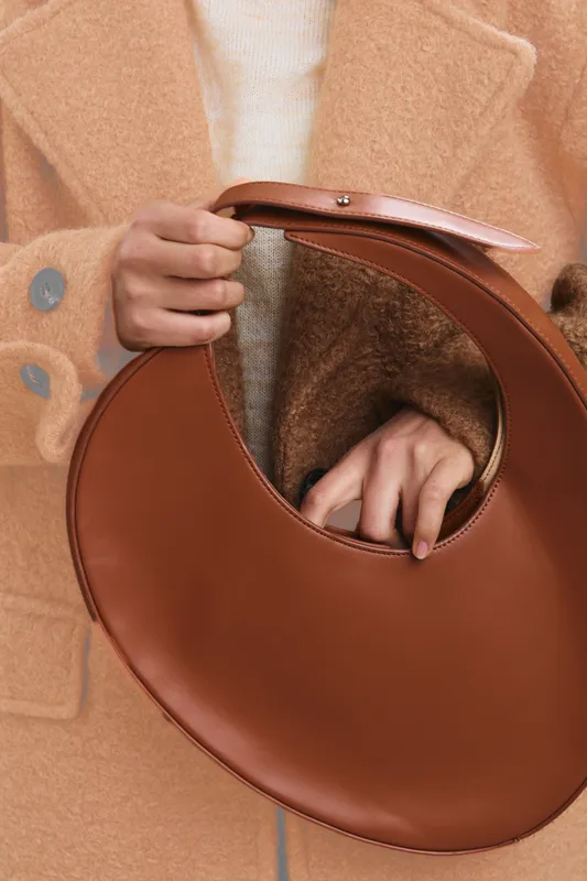 An asian woman wears a camel coat and holds a round brown handbag, presenting a stylish fashion moment with soft textures and neutral tones in a calm composition.