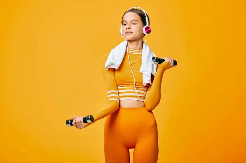 Female fitness model in bright orange sportswear holds dumbbells, with a towel and headphones, set against a vivid orange studio backdrop, showcasing energy, determination and an active workout