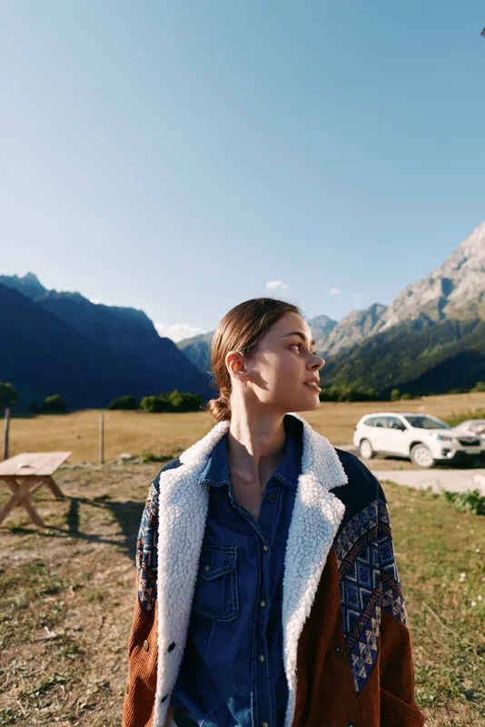 Woman portrait in mountains near parked car on meadow, wearing warm jacket and denim shirt, travel and nature scene with clear sky, outdoor lifestyle and peaceful scenery