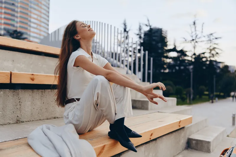 woman enjoying peaceful moment outdoors sitting on wooden steps in casual white outfit smiling with eyes closed KEY: woman, peaceful, outdoors, casual, white, smiling, relaxation, sitting, steps, joy,