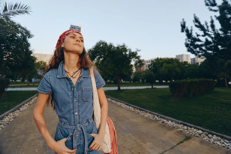 Lifestyle woman in modern boho-western denim street style with red bandana and layered necklaces, enjoying outdoor urban park, film color tones, casual confident pose.