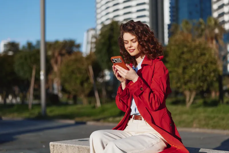 Young woman with curly hair and red jacket sits on a concrete bench outdoors while using her smartphone with a focused and content expression.