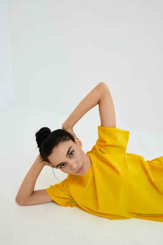 Young woman lying on white floor wearing bright yellow shirt, showing relaxed pose with hand behind head and calm facial expression.