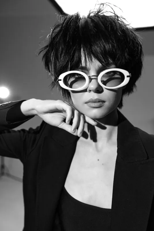 Black and white portrait of stylish woman wearing white sunglasses and black blazer with trendy short hair.