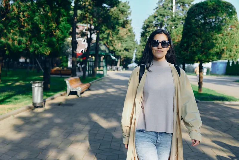 Woman walking in a park on a sunny day, wearing a beige coat, gray sweater and jeans, sunglasses, along a paved path with trees and benches, casual outdoor fashion moment