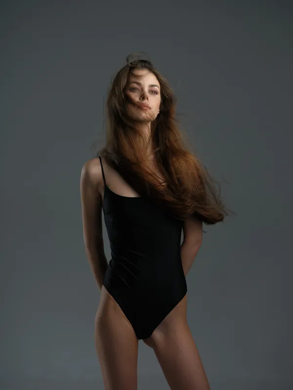 Young woman in black bodysuit with flowing hair standing against neutral gray background, natural beauty, fashion style.