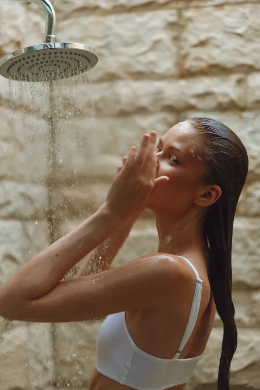 Woman showering outdoors under rainfall water, wearing white bra, wet hair slicked back, natural stone wall background, soft natural lighting, serene mood.