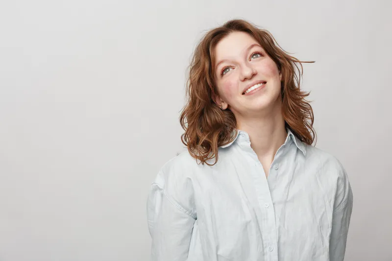 Young woman with red hair and light blue shirt looks up smiling on grey background. Concept of happiness, hope, casual style, and natural beauty in studio portrait.