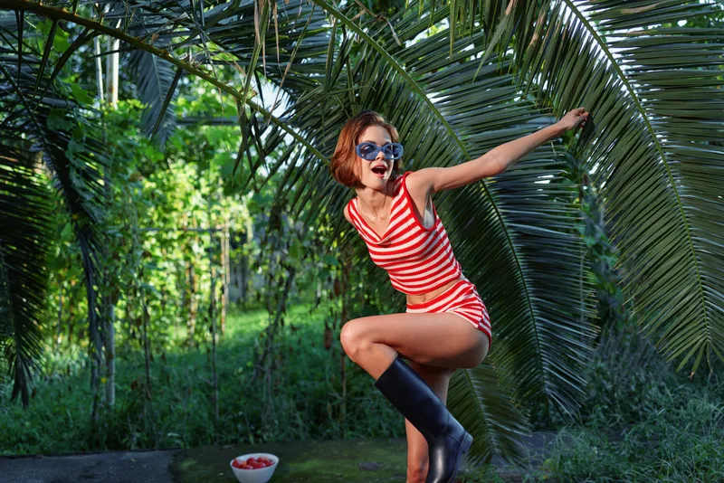 A playful woman wearing a red and white striped swimsuit poses beside large palm leaves in a sunny tropical setting. She wears sunglasses and boots, exuding bold summer energy.