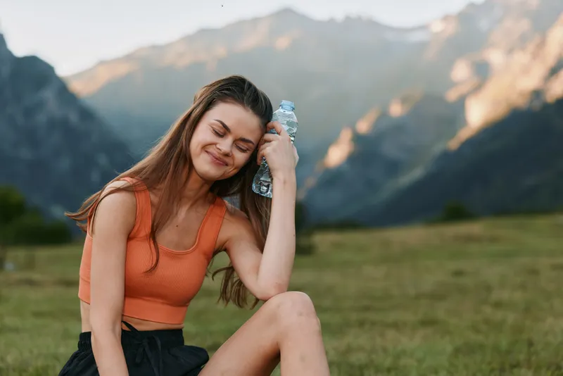A cheerful woman sits outdoors on a sunlit field, resting with a water bottle in hand, smiling softly as mountains rise in the background and the day feels peaceful.