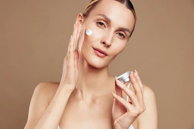 Woman beauty skincare cream face moisturizing portrait, young woman applies moisturizer with fingers, holds jar, fresh skin look, studio lighting, neutral background, high detail