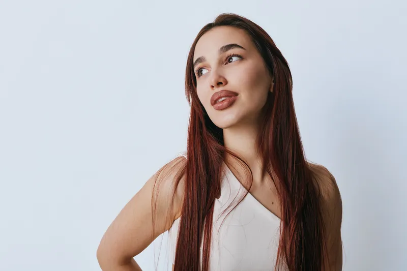 Woman with long red hair and natural makeup wearing white top posing against plain light background. Female model looking aside with confident expression and casual style in studio.