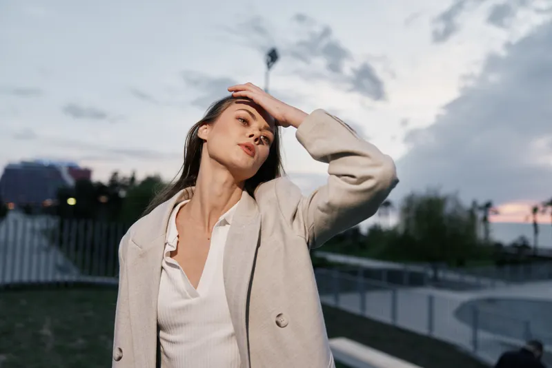 Confident businesswoman in a stylish light jacket poses outdoors at sunset, embodying modern professionalism and elegance, with a vibrant urban background.