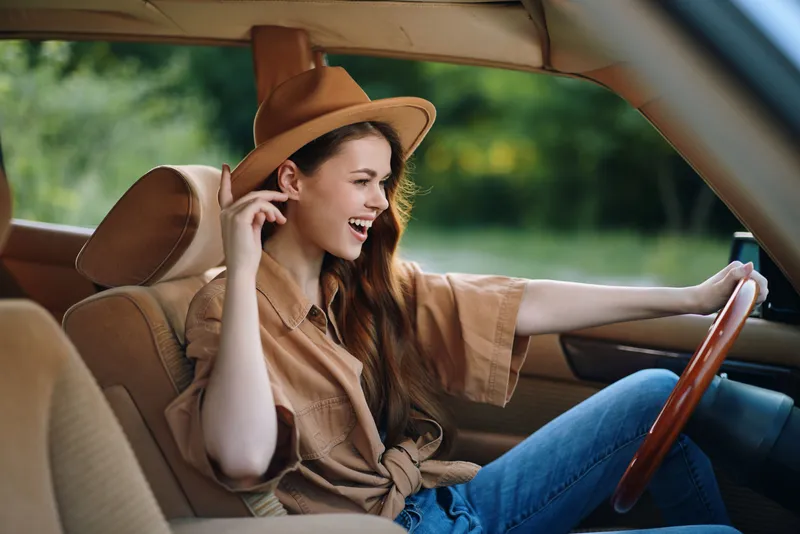 Cheerful young woman driving a vintage car, wearing a stylish hat and casual outfit, expressing joy in a vibrant outdoor setting, summer vibes.
