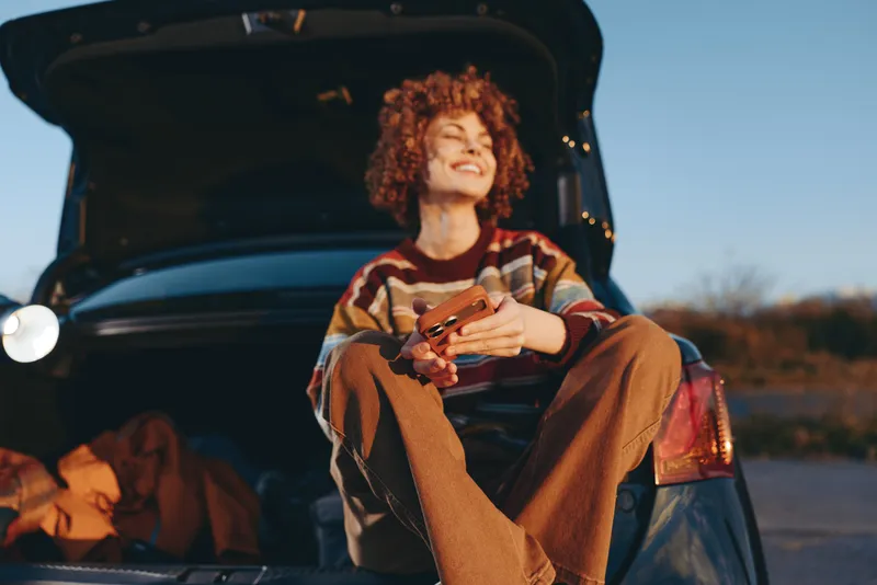 Woman lifestyle photo, curly hair, sitting on open car trunk outdoors at evening, holding a smartphone, relaxed smile with sparks and warm glow, casual striped shirt, dusk atmosphere.