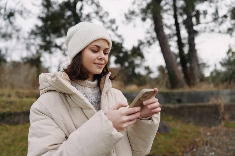 Casual woman in a warm coat and beanie using a smartphone outdoors in a park during chilly weather, enjoying a tech moment and fresh air.