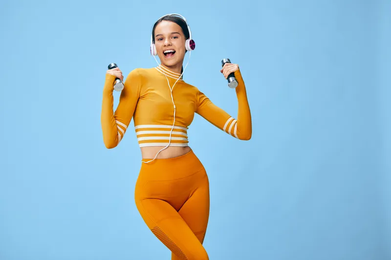 Female fitness enthusiast in orange activewear holds dumbbells against a bright blue backdrop, showcasing energy, strength and motivation during a dynamic workout in studio.