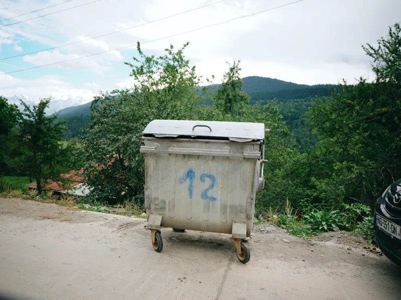 Dumpster on a rural roadside with hills and trees, weathered container, metal lid, wheels, blue spray painted number twelve, gravel path, overcast sky, distant mountains, calm outdoor waste storage