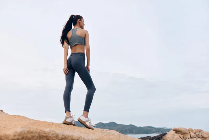 Confident young woman in activewear enjoying a scenic view by the sea, emphasizing fitness and lifestyle, wearing gray leggings and sports top