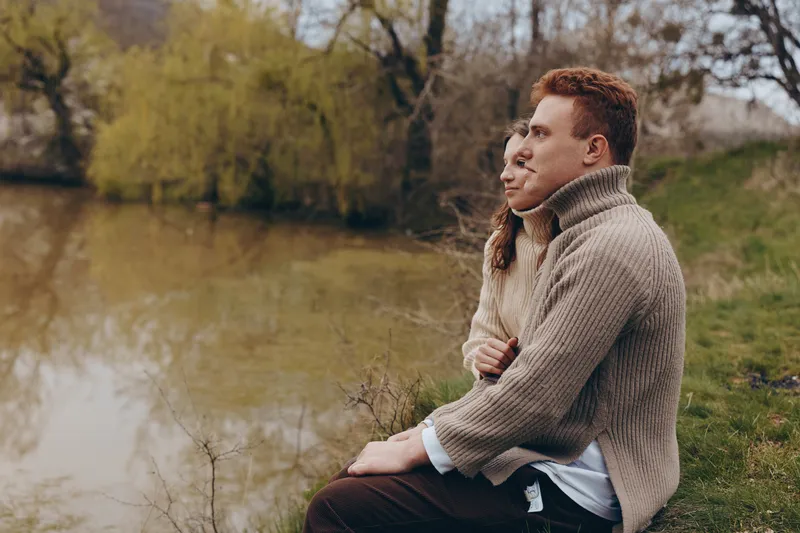A couple sits close on a bench by a calm pond, embracing in a cozy sweater. A peaceful lifestyle moment captured outdoors, highlighting connection, warmth, and quiet reflection.