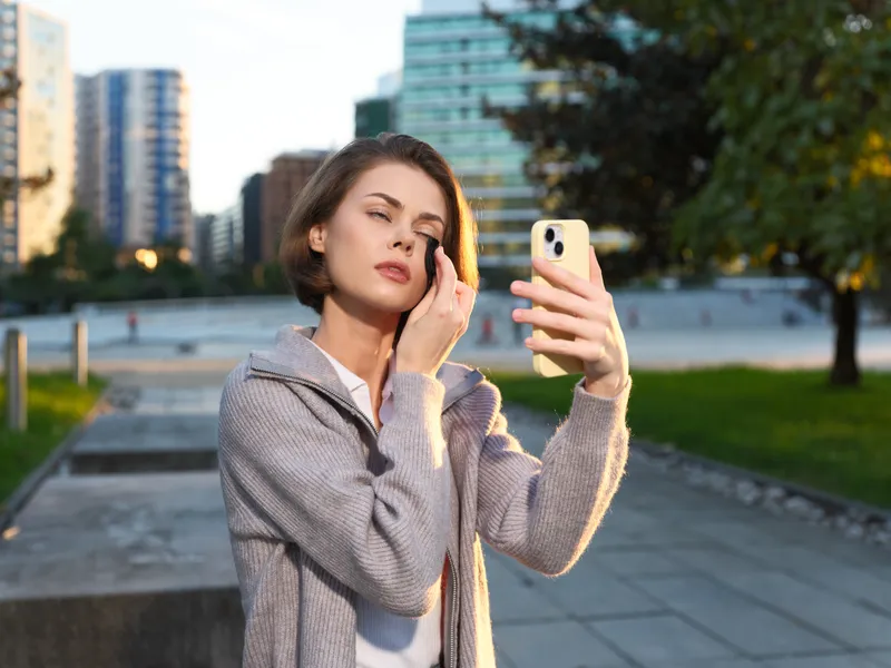 A thoughtful woman takes a selfie with a smartphone in an urban park setting. She wears a light coat and a calm smile, while modern buildings rise behind her, creating a dynamic city vibe.