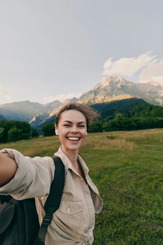 Cheerful woman outdoors, smiling into the camera, backpack ready for a hike, wide green meadow with distant mountains, soft light, natural colors, relaxed lifestyle moment today