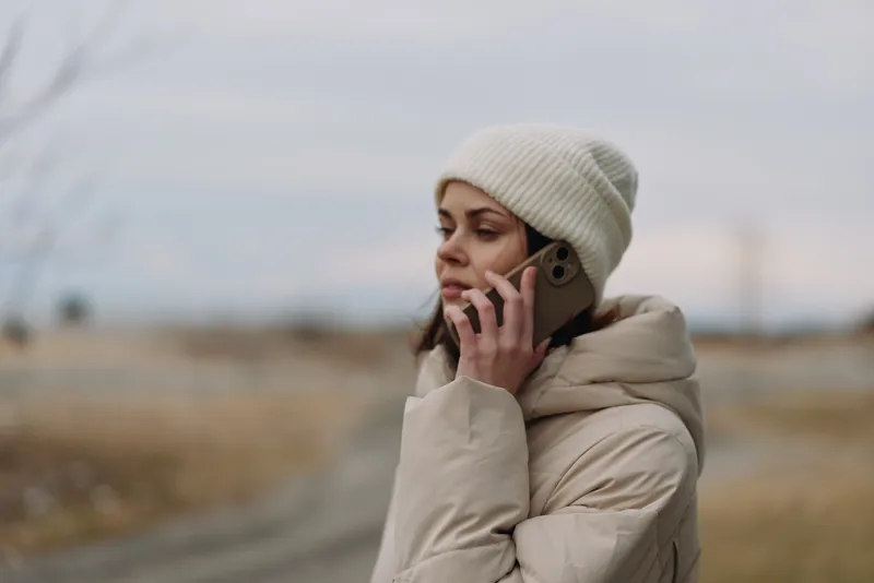 Winter scene with a woman in a warm coat and beanie talking on a cellphone outdoors, calm landscape and soft light creating a quiet moment of connection.