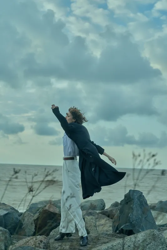 Figure standing on rocky shoreline with flowing coat, wind-swept pose by the sea, dramatic clouds overhead, conveying freedom, movement, contemplation and solitude in ocean landscape