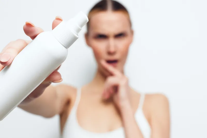 Young woman holds white spray bottle close to camera with blurred face in background, shows skincare product, beauty concept, isolated on white background.