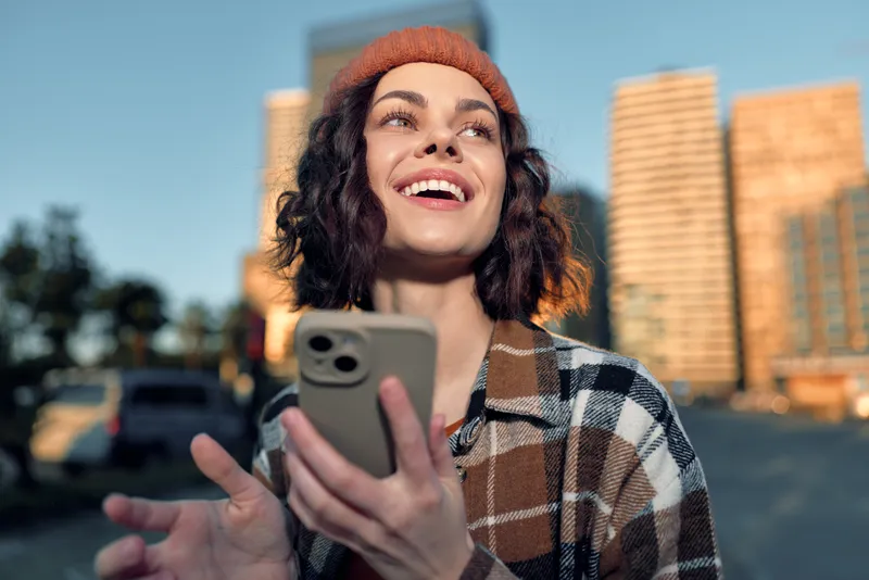 Woman with smartphone smile, smiling in urban street at golden hour glow, candid authenticity and lifestyle portrait; beanie, happiness, mindful living and emotional storytelling capture modern city