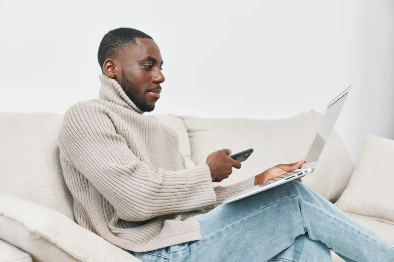 Young Black man in a cozy beige sweater interacting with his smartphone while working on a laptop in a modern living room setting, showcasing a relaxed vibe