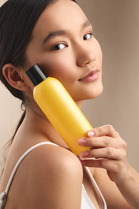 Asian woman is photographed in a close up studio portrait, holding a yellow bottle near her shoulder, highlighting beauty and haircare product with soft lighting and a clean, modern feel.