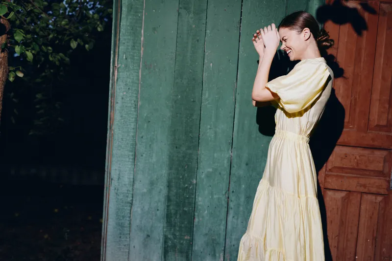Woman in yellow dress standing by green door in sunlight smiling and shielding face, authentic inclusive portrait with natural light vintage outdoor lifestyle