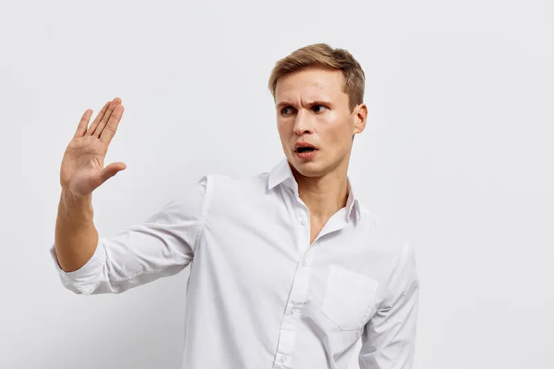 Confused man in white shirt looking surprised and raising his hand, studio portrait of puzzled young male expressing confusion or shock, isolated on plain light gray background, people emotion concept