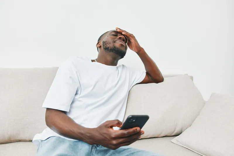 A stressed young African American man sitting on a couch, holding a phone and covering his forehead, depicting frustration and anxiety in a minimalist setting