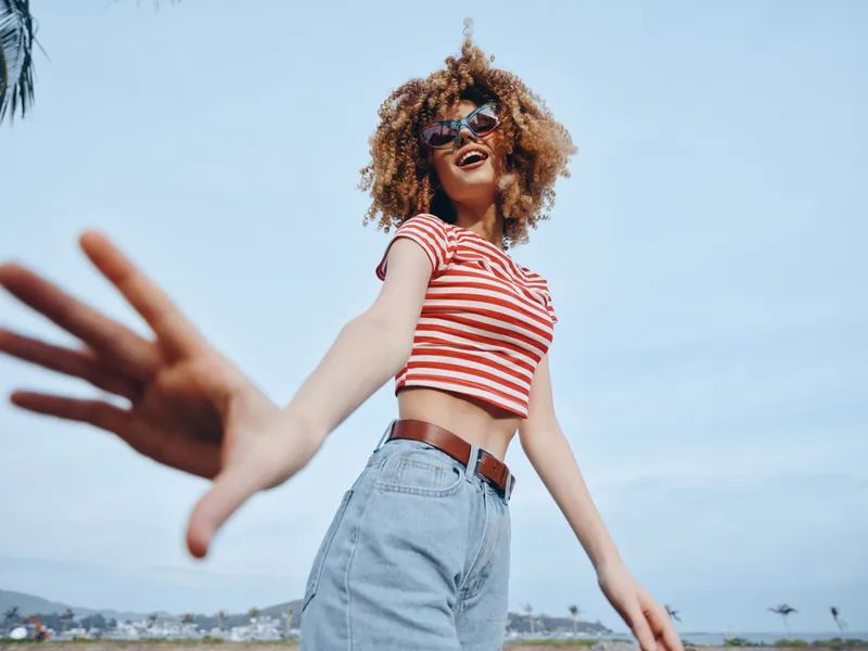 A joyful woman with curly hair wearing a red striped crop top and light denim jeans stands outdoors by the coast, reaching playfully toward the camera.
