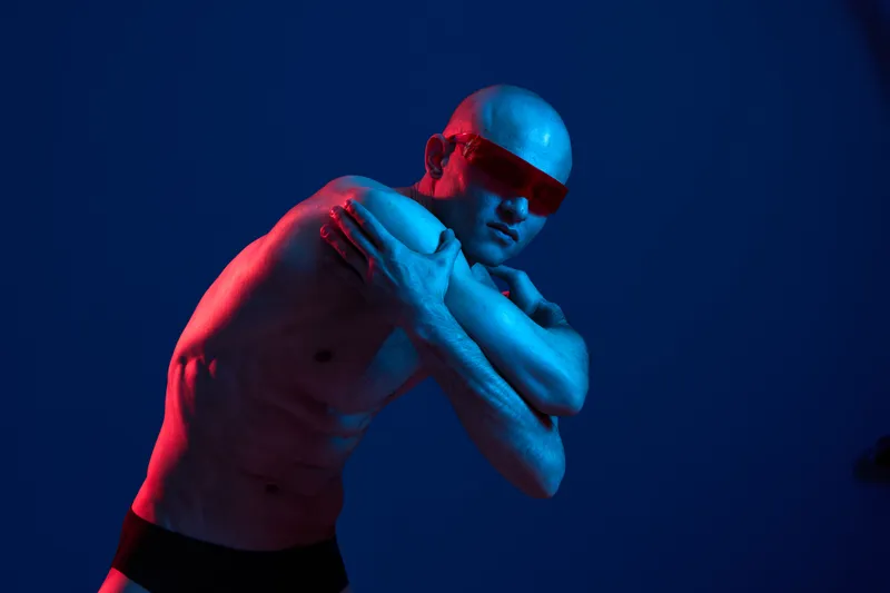 Athletic man striking a unique pose in a vibrant blue and red light, showcasing strong, muscular features and a contemplative expression. Perfect for fitness themes.