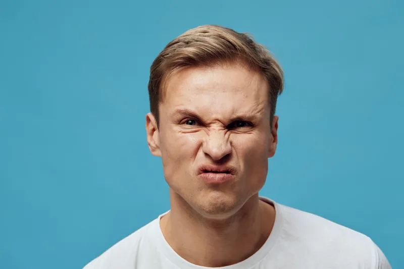 Close-up of young caucasian man making a scrunched face, showing irritation or disapproval, wearing a white t-shirt against a solid light blue background. People emotions concept.