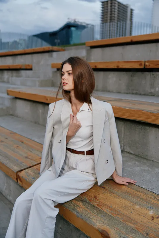 Fashion woman wearing beige blazer and pants sitting on wooden steps in urban outdoor setting with modern city background and natural daylight