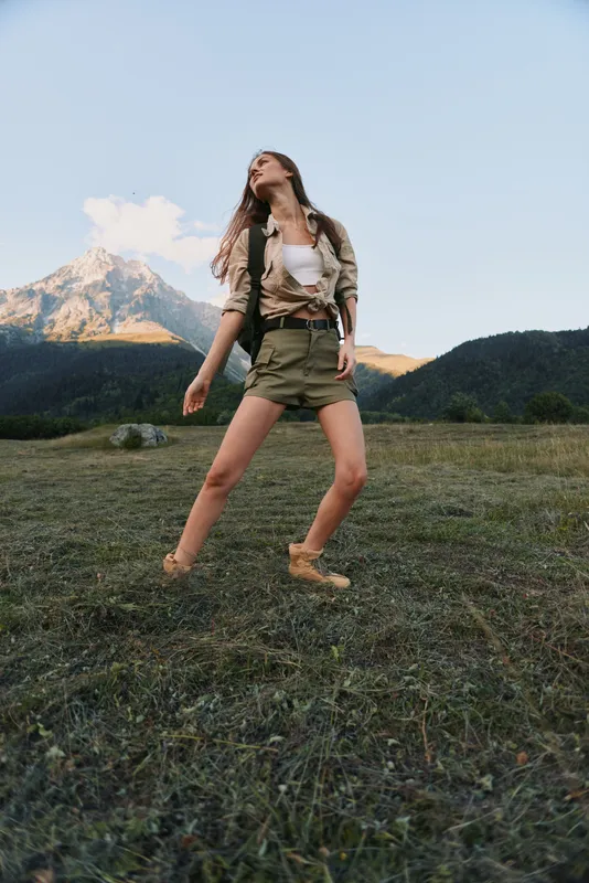 Relaxed woman in a grassy meadow explores an outdoor landscape, wearing casual attire. Bright natural light, expansive sky, and distant mountains convey freedom, nature, and lifestyle.