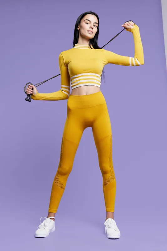Young woman sport fitness wearing yellow athletic outfit with resistance band, posing confidently against purple background in activewear for strength training.