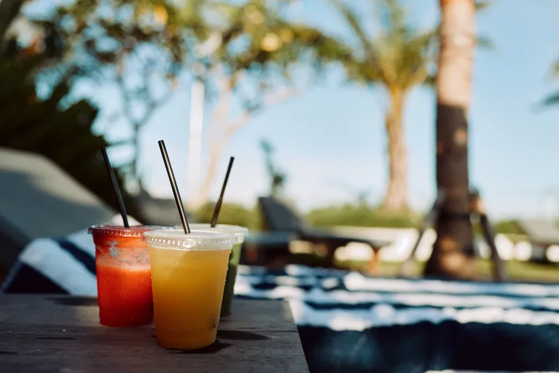 Two cold summer drinks in plastic cups with straws stand on a wooden table near a pool with palm trees in the background on a sunny day.