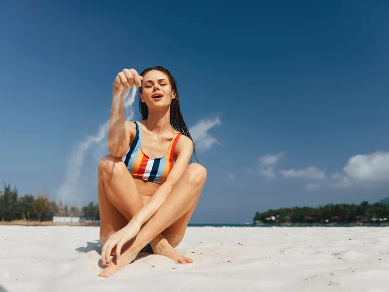Female sits on white sand at the beach, wearing a multicolor striped bikini, sunlit skin and relaxed smile, toes buried in sand, clear blue sky overhead, capturing carefree summer energy.