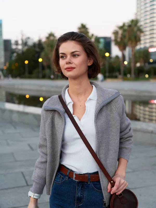 A confident woman stands outdoors in a modern city plaza, wearing a coat and casual outfit, smiling softly as she looks ahead with a calm, focused attitude.