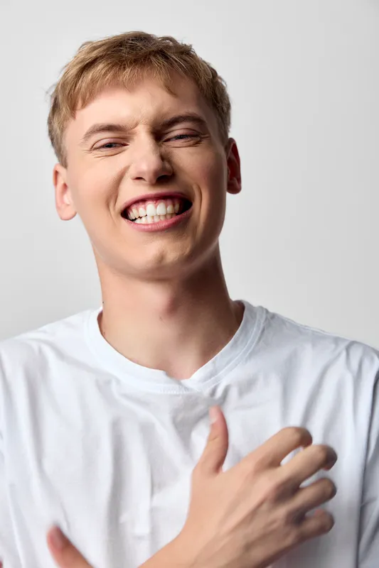Smiling man in a white tshirt expresses joy and cheer, hand over chest in a candid studio shot, capturing happiness, energy and confidence for lifestyle, wellness and positive mood themes