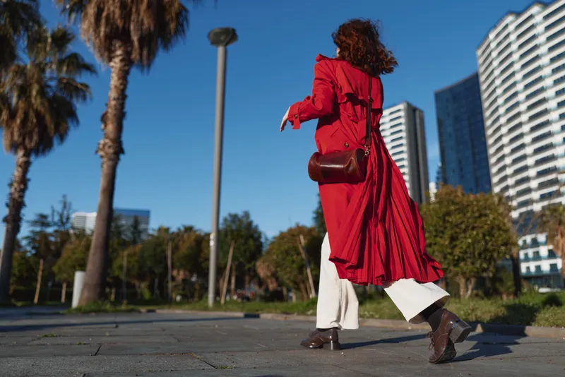 Young woman in a red coat walks through a sunny urban park with palm trees and tall buildings, expressing confident motion under a clear blue sky, a light breeze