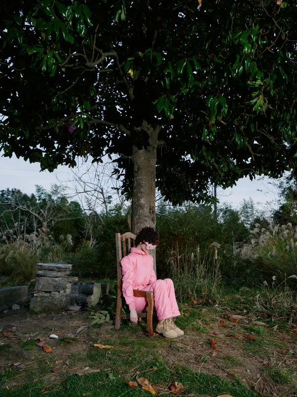 Person sitting on a wooden chair under a large leafy tree in a quiet garden, wearing a pink outfit, surrounded by grass, leaves, and distant hedges, with soft afternoon light and a peaceful rural