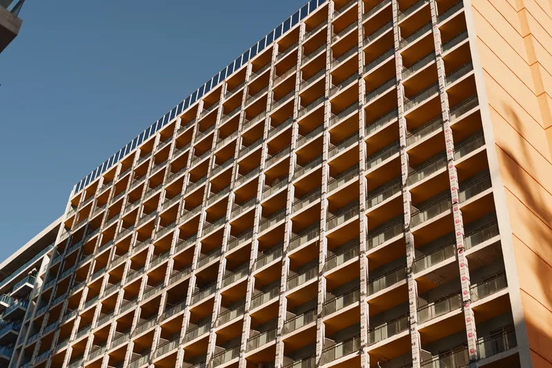 Sunlight casts sharp shadows on the facade of a modern apartment building with repetitive balconies under a clear blue sky.