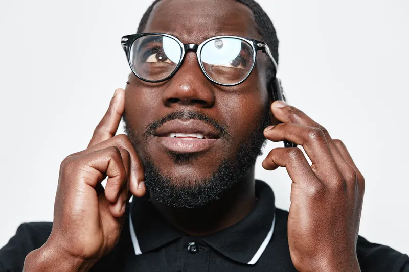 African American man with dark skin wearing glasses and black polo shirt talking on smartphone with thoughtful expression isolated on white background.