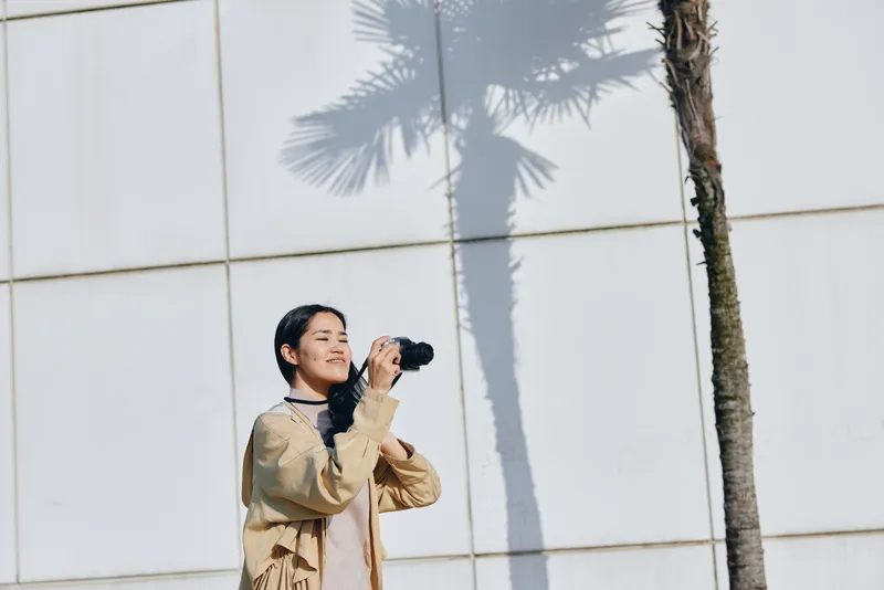 Woman photographer outdoors beside a modern wall, holding a camera to her eye as palm shadows stretch across the surface, bright daylight framing a candid travel moment